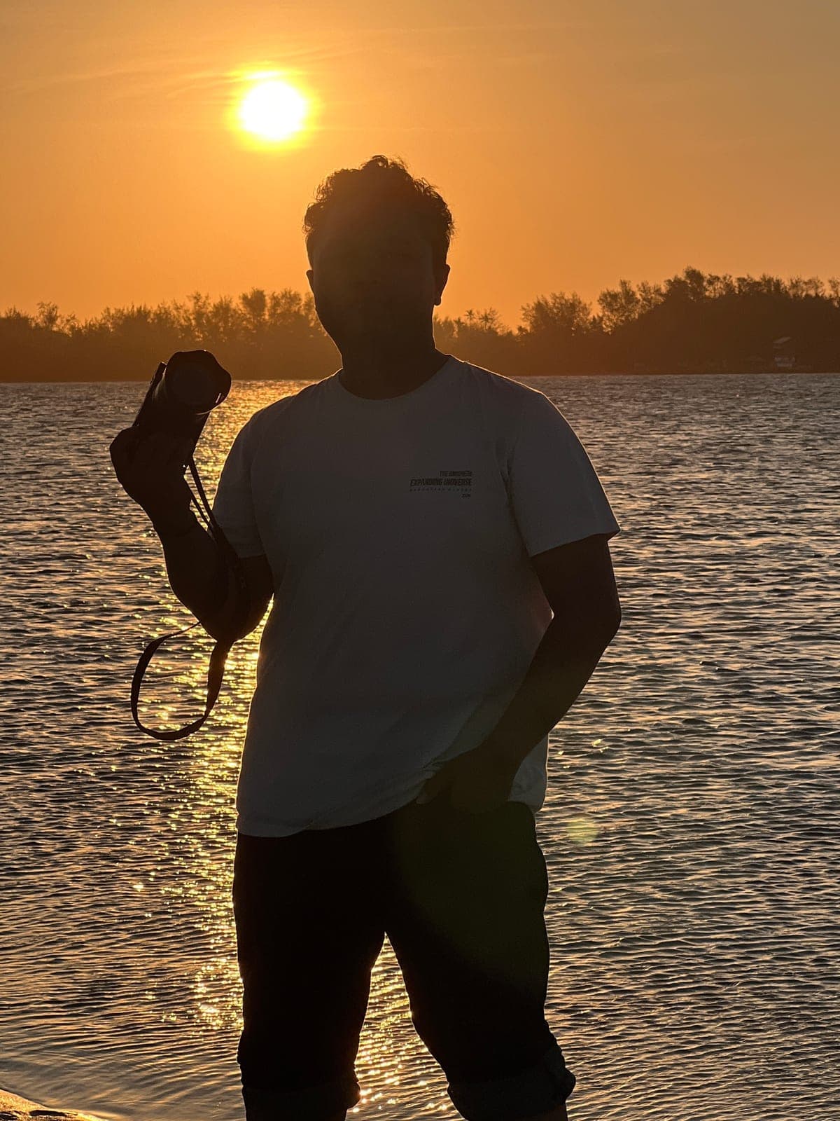 Farrel memegang kamera di tepi pantai saat matahari terbenam
